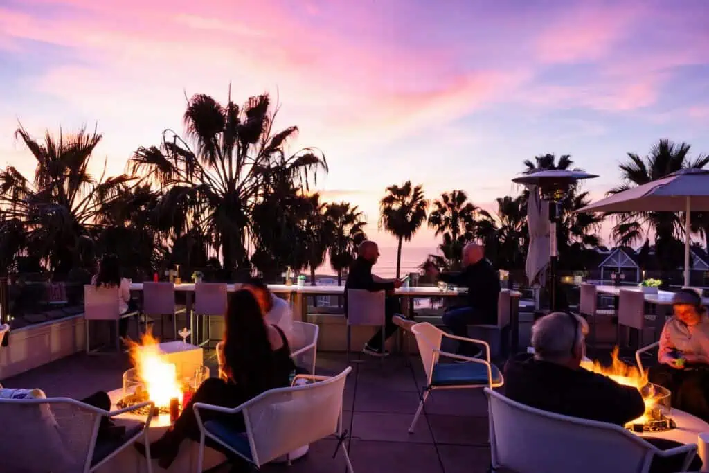 People sit around fire pits and tables on an outdoor patio at sunset, with palm trees and the ocean visible in the background.