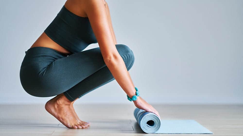 A person in athletic wear squats while rolling up a yoga mat on a light-colored floor, embracing longevity trends Seattle is known for.