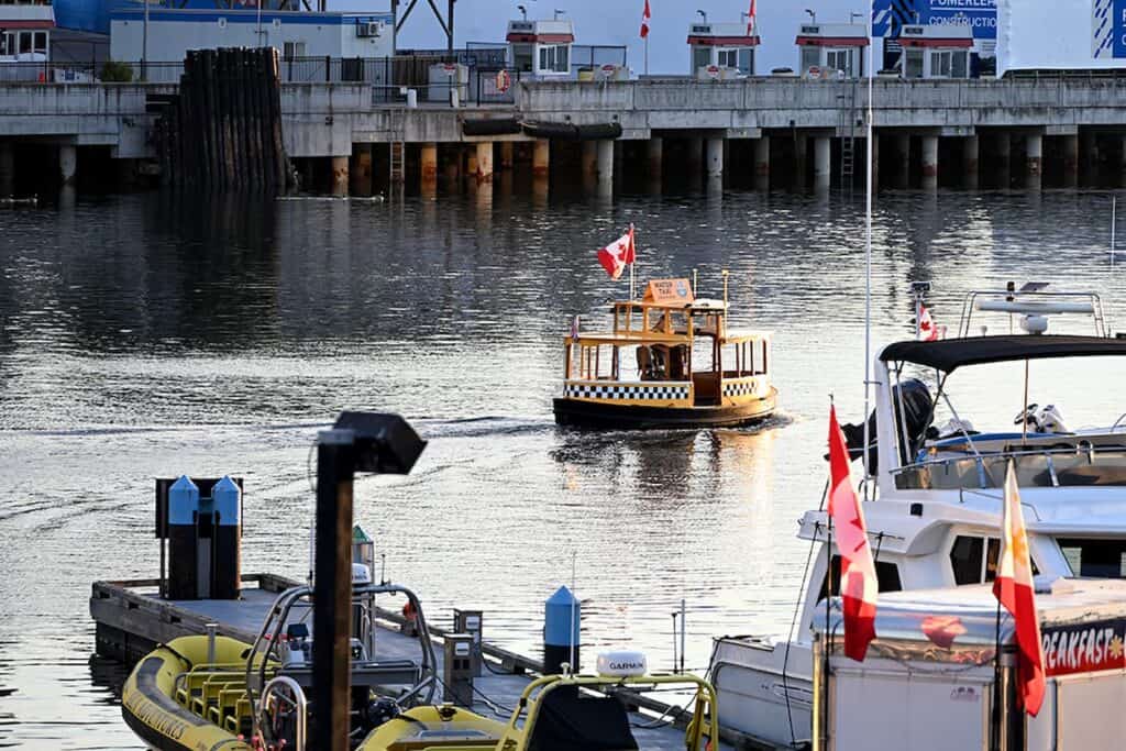 A small water taxi with a Canadian flag approaches a dock area with several boats, reflecting on calm water near an urban waterfront.