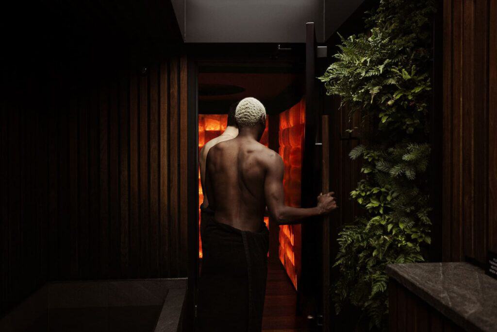 A person with a towel wrapped around their waist opens a door to a dimly lit room with glowing orange walls, surrounded by wooden and plant-covered walls.
