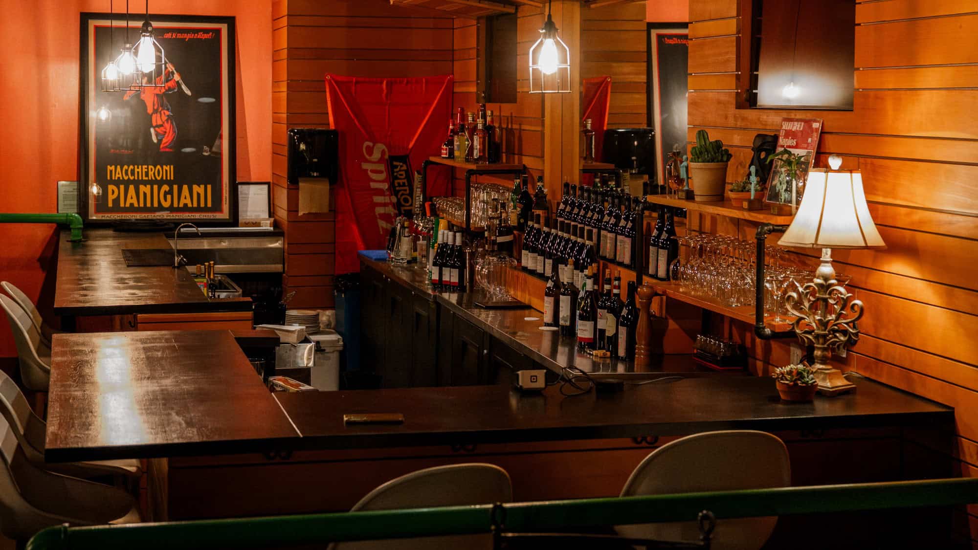 A cozy bar with wooden walls, shelves of wine bottles, bar stools, glasses, a decorative lamp, and a vintage poster on the wall.