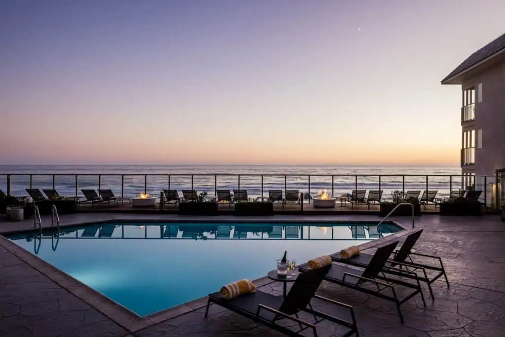 Outdoor pool area with lounge chairs and towels overlooks the ocean at sunset, with a clear sky and calm water, next to a light-colored building.
