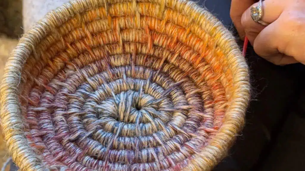 A hand holds a partially finished, coiled basket woven with multicolored yarn in concentric rings.