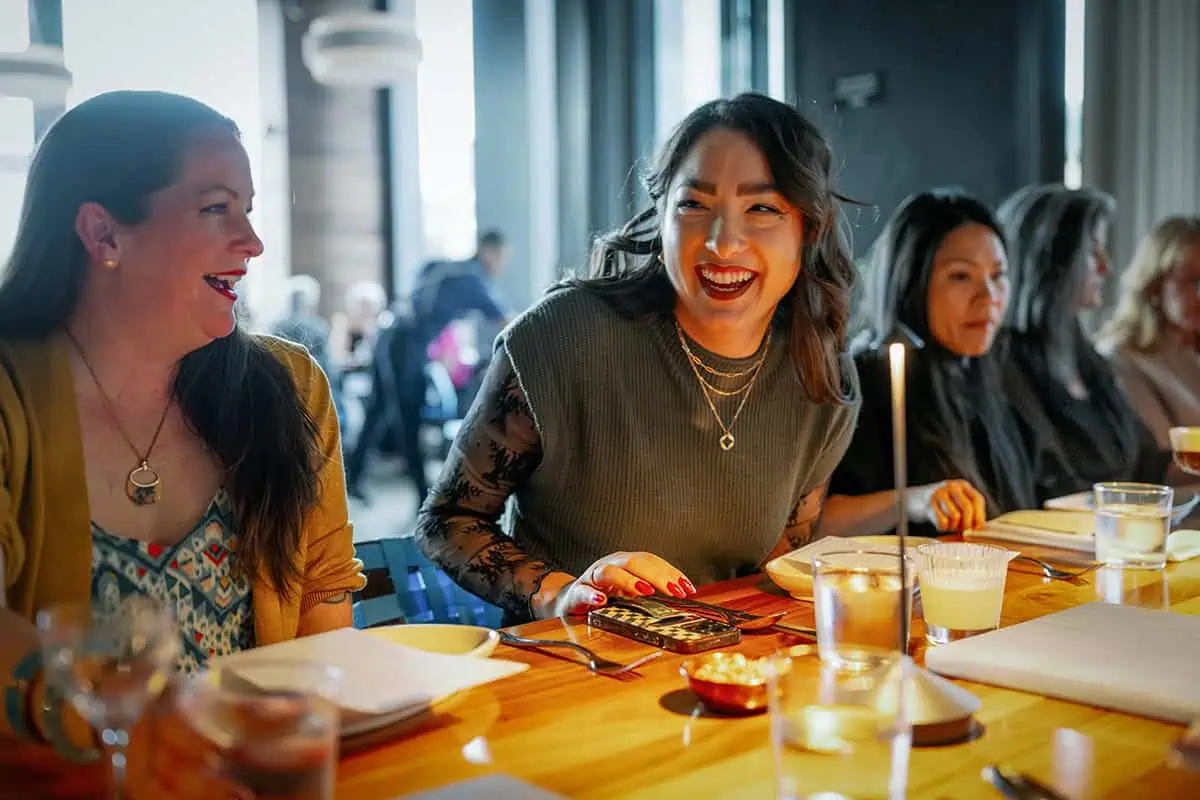 Women sit at a restaurant table in Seattle, talking and laughing, with menus, water glasses, and candles on the wooden table—making it the perfect spot for event planning or meeting with trusted PNW vendors.