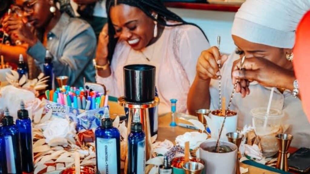 Several women sit at a table making Noir Lux candles, surrounded by candle supplies, essential oil bottles, and colorful markers as they experiment with unique fragrance blends.