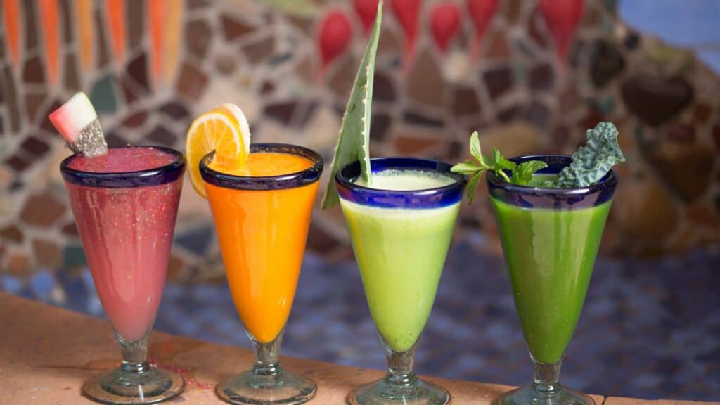Four tall glasses with colorful smoothies—pink, orange, light green, and dark green—each garnished with fruit or greens, displayed on a counter against a mosaic background.