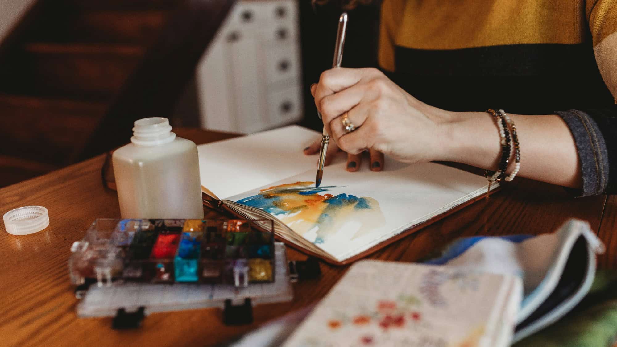 A person uses a paintbrush to create a watercolor painting in a sketchbook on a wooden table, with watercolor paints and a jar nearby.