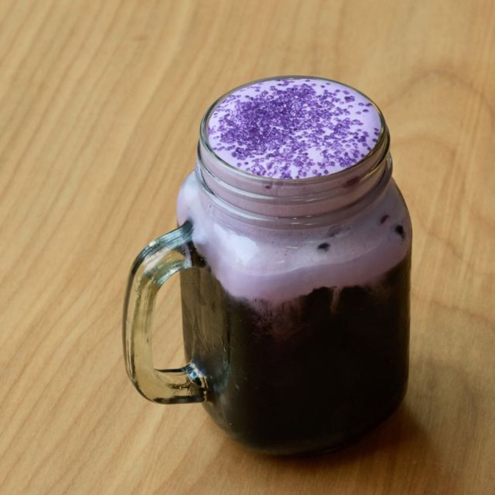 Mason jar filled with a dark iced drink, topped with purple foam and purple sprinkles, on a light wood surface—an eye-catching creation inspired by the next generation coffee served at Seattle coffee shops.