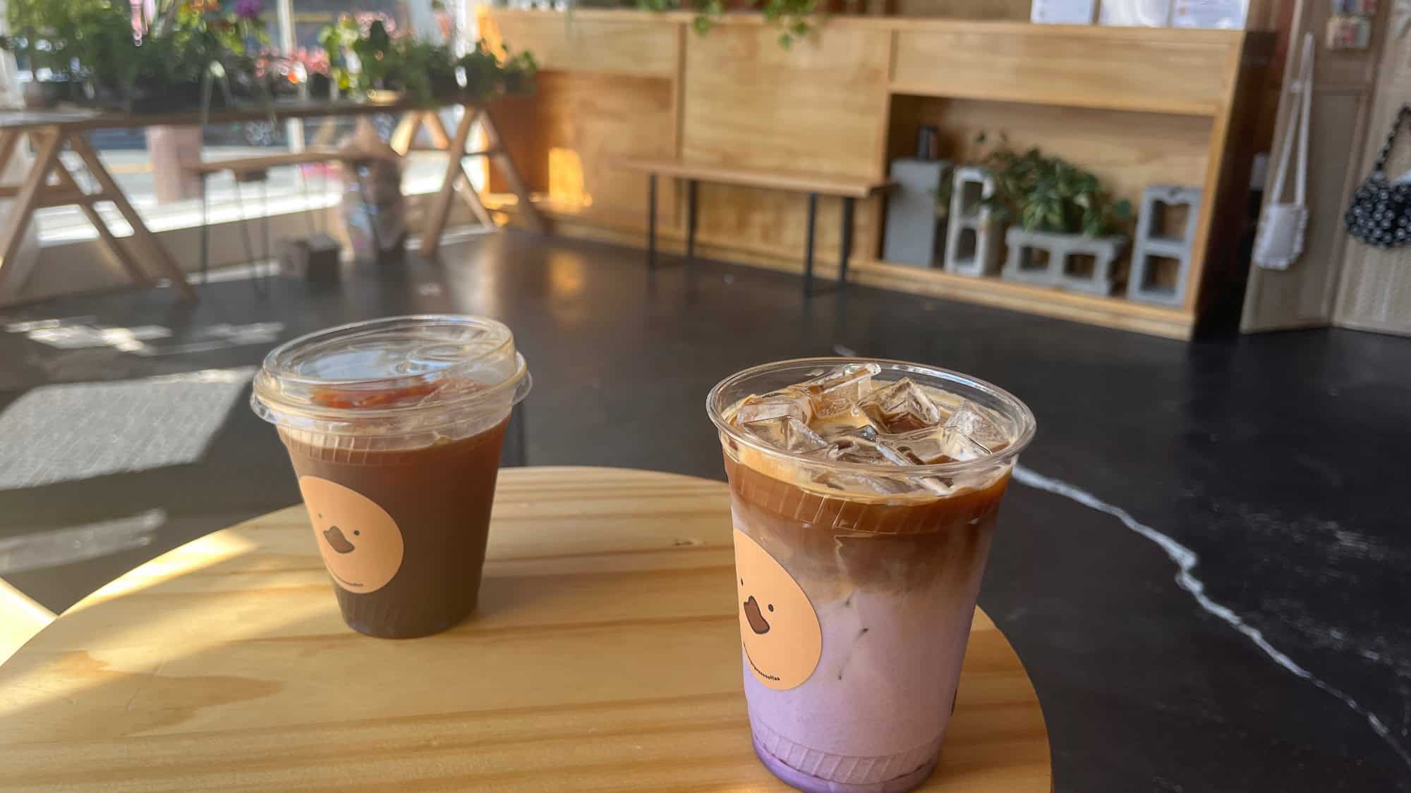 Two plastic cups of iced coffee drinks on a round wooden table in a cafe with plants and wooden shelving in the background.