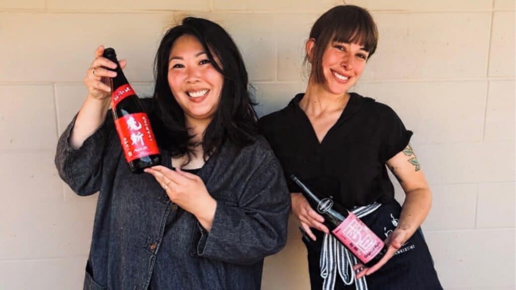 Two women stand side by side smiling, each holding a bottle with a pink or red label in front of a plain light-colored wall.