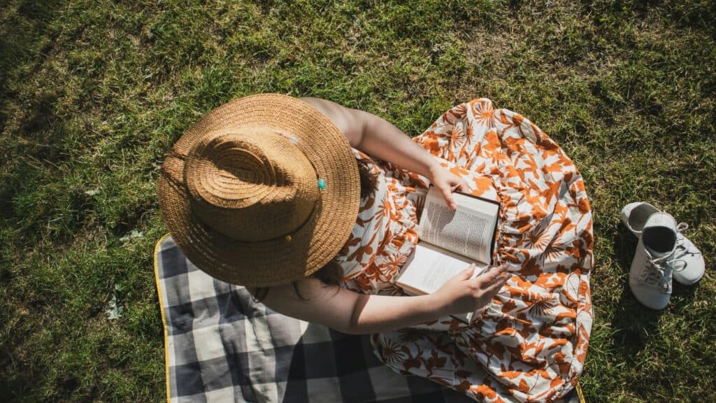 Person in a straw hat and patterned dress sits on a blanket reading a book on grass—a perfect example of stress-free activities ideal for PNW daytrips; shoes are placed nearby.