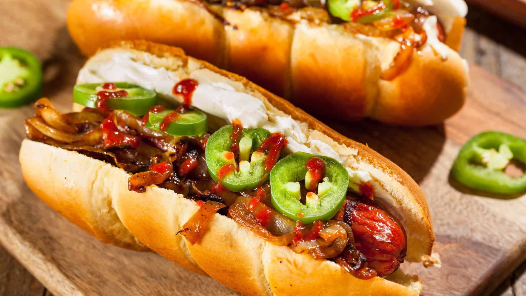 Two hot dogs topped with sliced jalapeños, grilled onions, and a drizzle of hot sauce channel the classic Seattle Dog vibes—worthy of any Restaurant Roundup—served on a wooden board.
