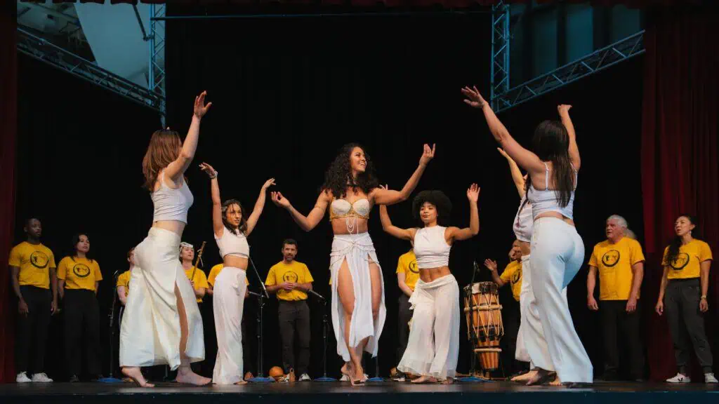 A group of women in white dance on stage with raised arms as people in yellow shirts and musicians with drums stand in the background.