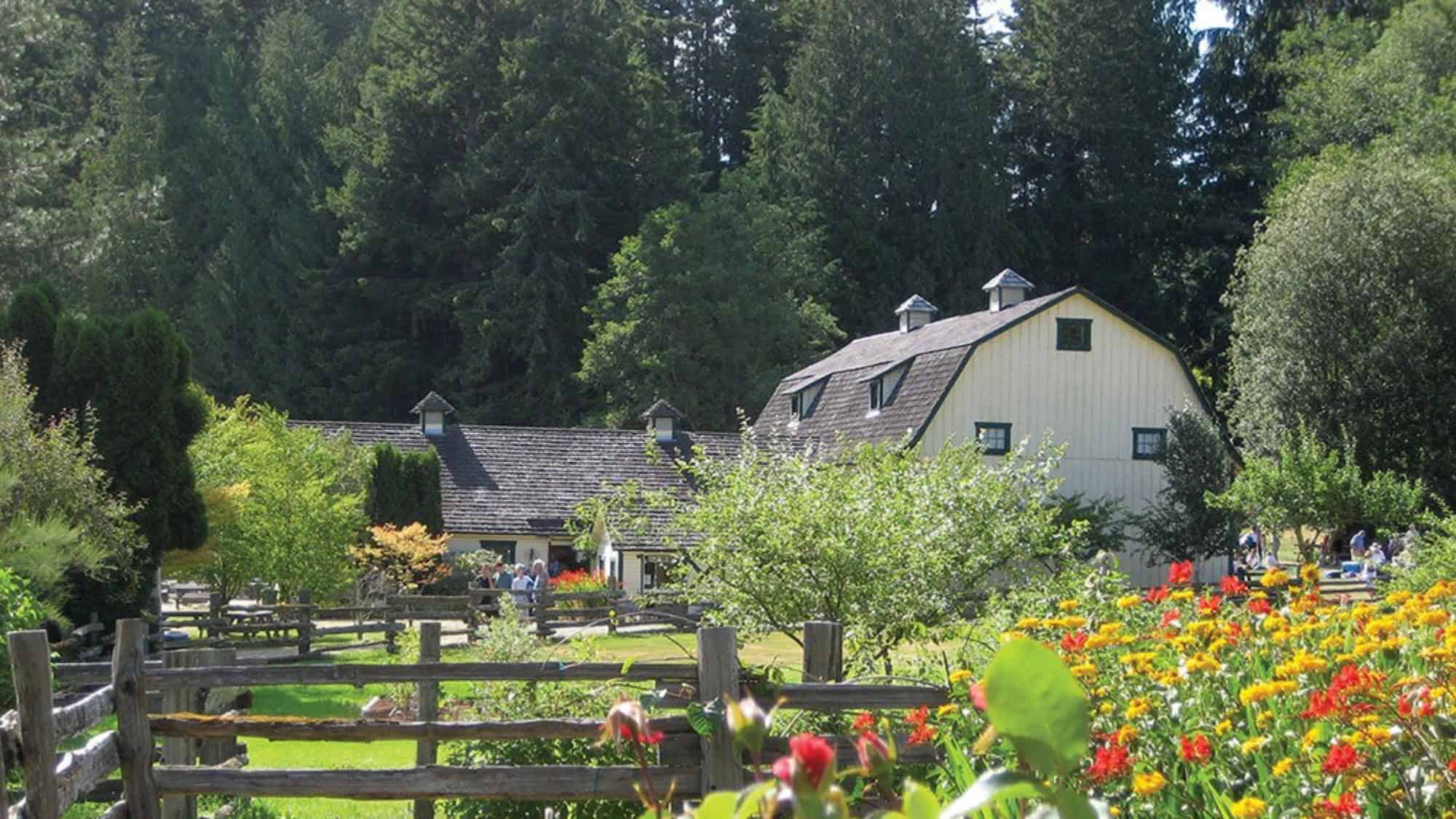 A white barn surrounded by trees, a wooden fence, and colorful flowers, with people walking and relaxing in the garden on a sunny day.