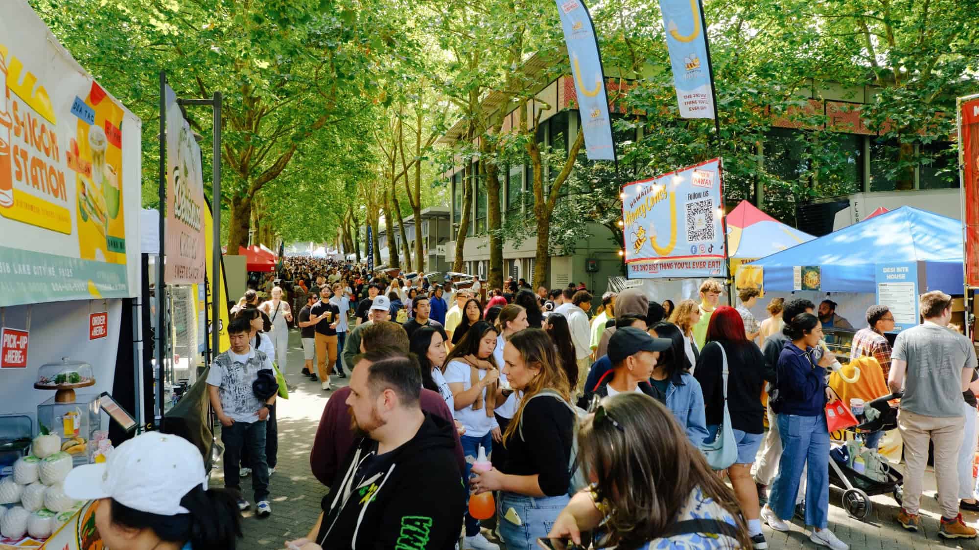 Crowd of people walking and browsing food stalls at an outdoor market lined with trees and colorful vendor tents during the day, reflecting the vibrant hospitality community coming together in support after tragedy.