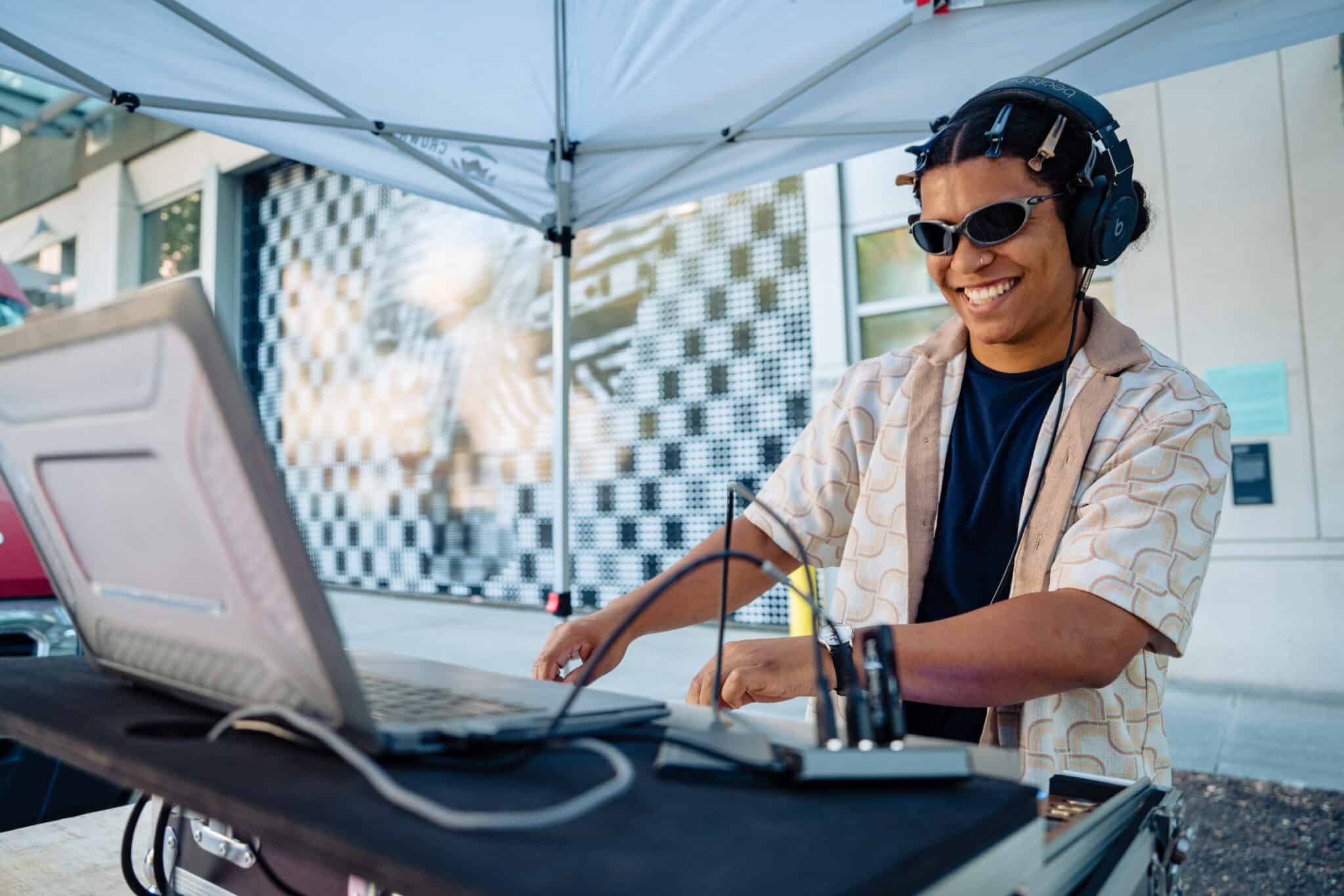 Person wearing headphones and sunglasses smiles while operating DJ equipment under a canopy outdoors, with a laptop and audio gear in front of them near Pine and Pike, just steps away from the Seattle Convention Center—where local food vendors add to the vibrant atmosphere.