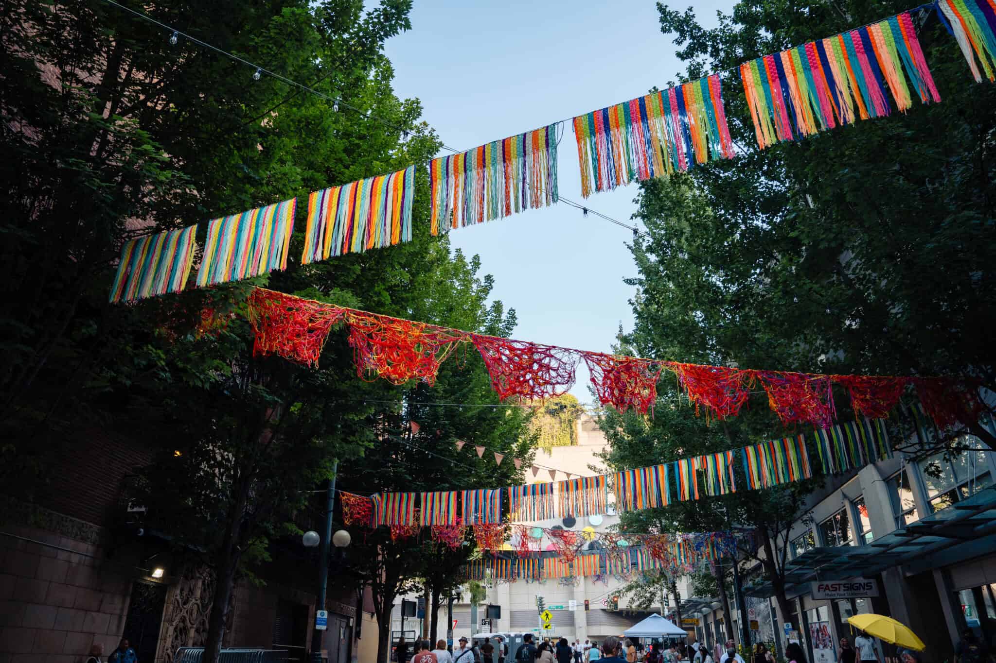 Colorful ribbons and red crocheted banners hang above Pine and Pike, where 9th Ave. bustles with people, trees lining the lively street. In the background, local food vendors set up tents, adding to the vibrant city scene.