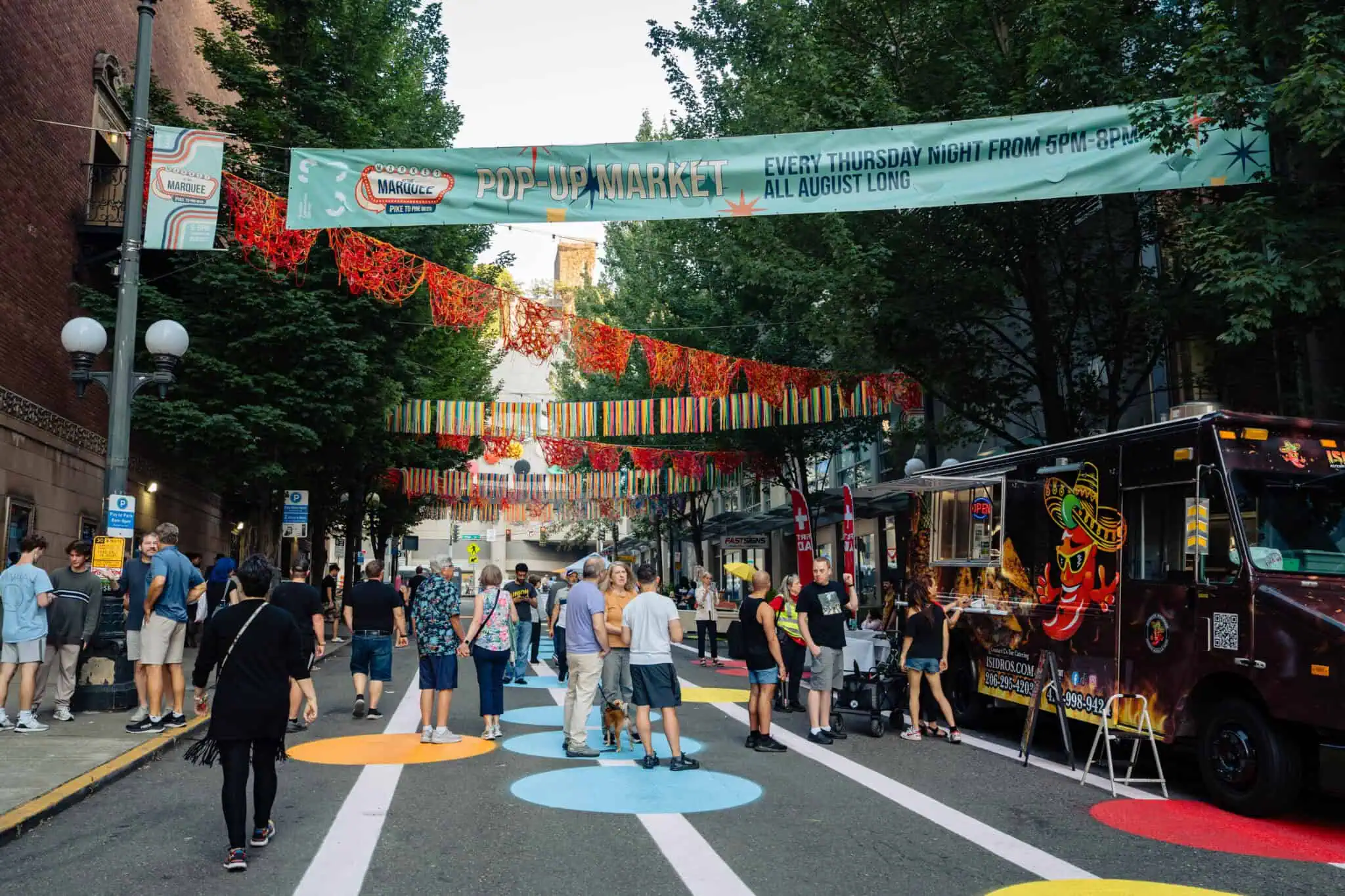 People gather at Pine and Pike for the lively Market at the Marquee, a street pop-up market adorned with colorful decorations, banners, and a bustling food truck. Painted circles mark 9th Ave, while trees line the street and buildings rise in the background.
