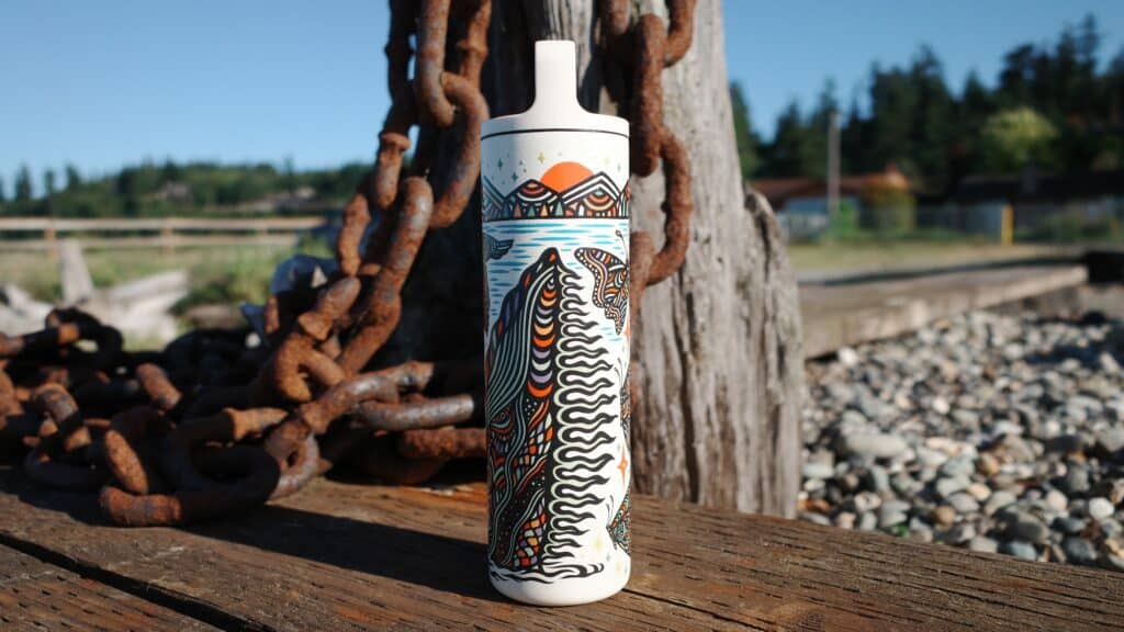 A ceramic bottle with intricate painted designs stands on a wooden surface near rusted chains, pebbles, and trees in the background.