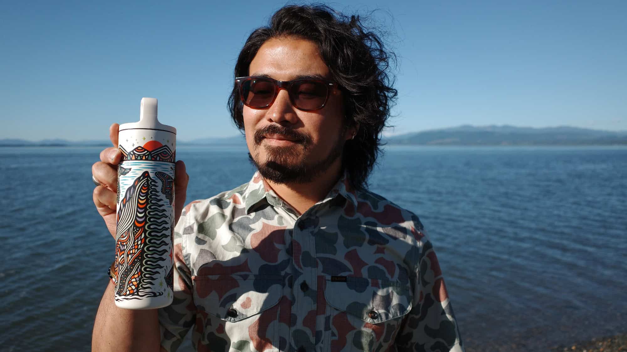 A man wearing sunglasses and a patterned shirt stands by a body of water, holding up a decorated white bottle with a colorful abstract design.