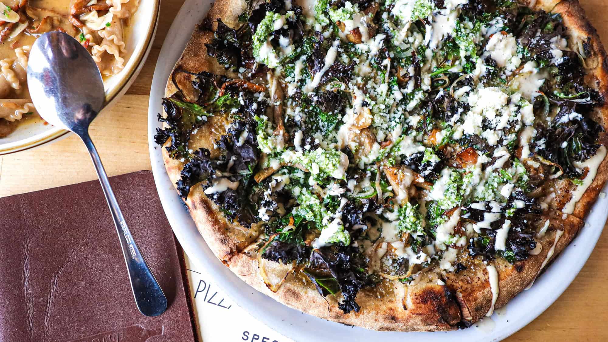 A close-up of a baked pizza topped with greens, cheese, and sauce, next to a pasta dish and a brown menu on a wooden table.