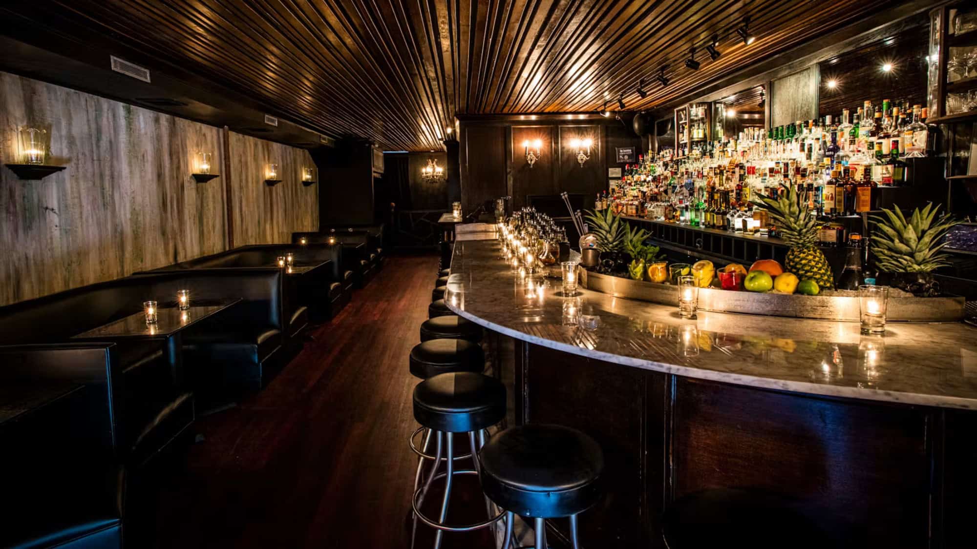 Dimly lit bar with a marble counter, barstools, fruit display, shelves of liquor bottles, and black leather booths along the wall. Candles provide soft lighting throughout the space.
