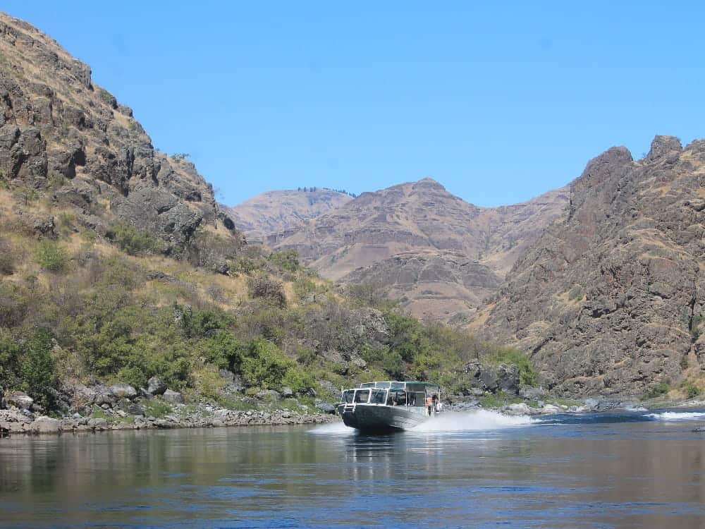 A motorboat speeds along a calm river, promising adventure as it weaves through rocky hills and sparse vegetation under a clear blue sky—an experience that captures the spirit of local culture.