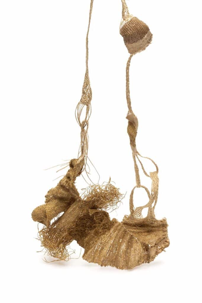 A delicate, gold-brown woven fiber sculpture with irregular shapes and frayed edges hangs from two twisted strands against a white background.