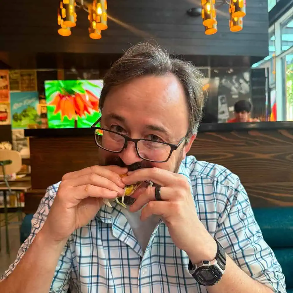 A man in glasses and a plaid shirt enjoys a sandwich made with local grass-fed beef at a restaurant, with a television and hanging lights in the background.