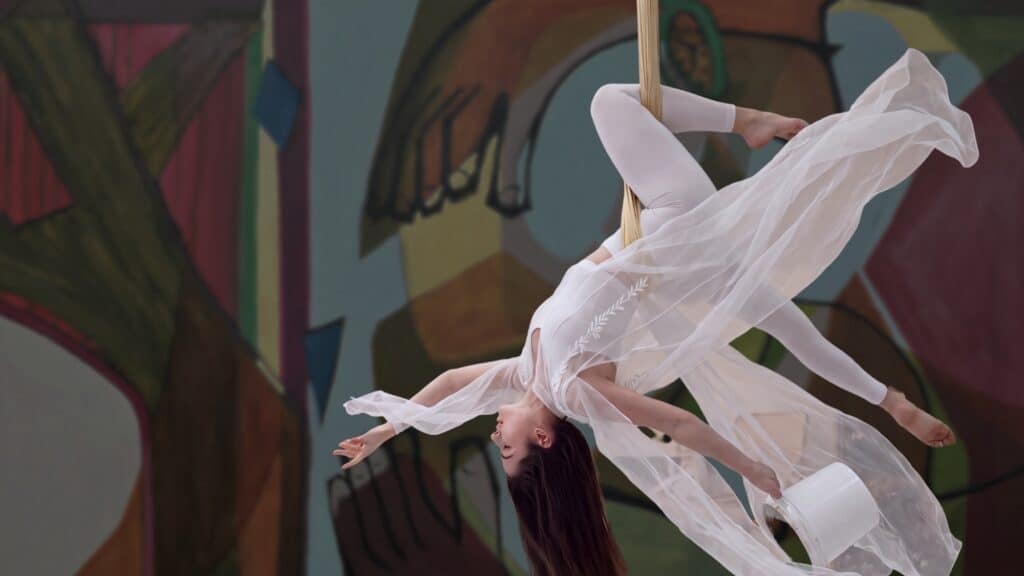 Aerial performer in a white costume hangs upside down on a rope, with sheer fabric flowing, against a colorful abstract mural background.