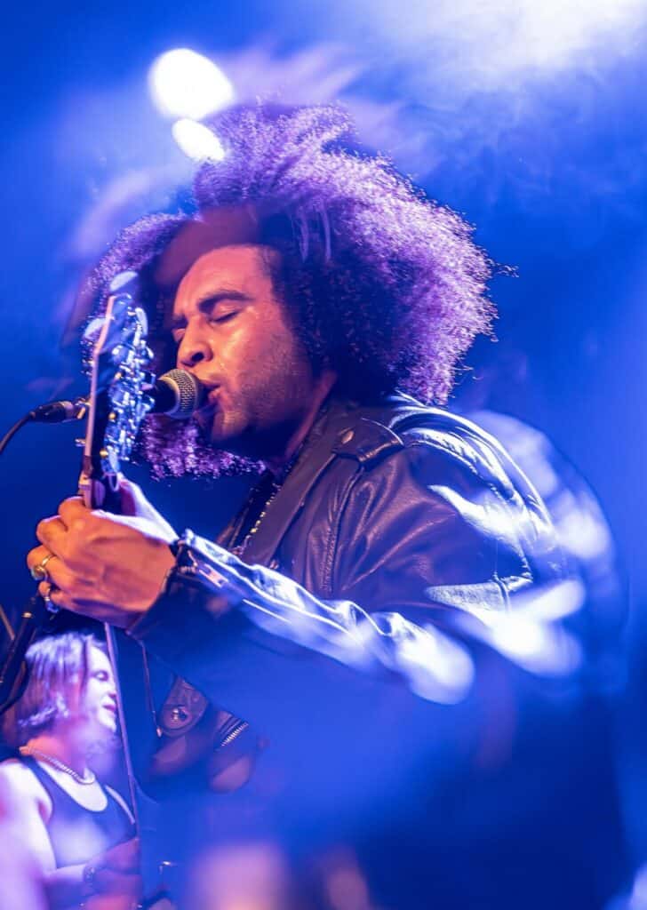 Musician with curly hair and leather jacket sings and plays guitar on stage under bright lights; another performer is visible in the background.