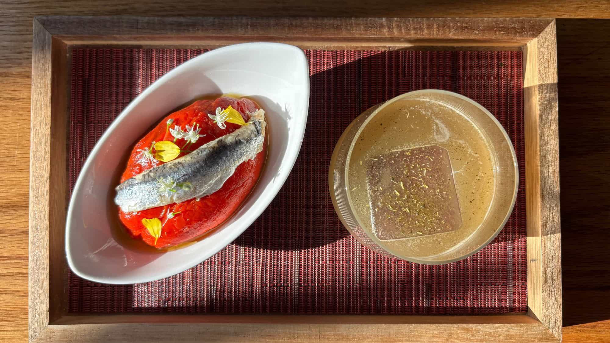 A wooden tray holds a white dish with a tomato topped with a small fish and herbs, next to a glass of light brown drink with a large ice cube.