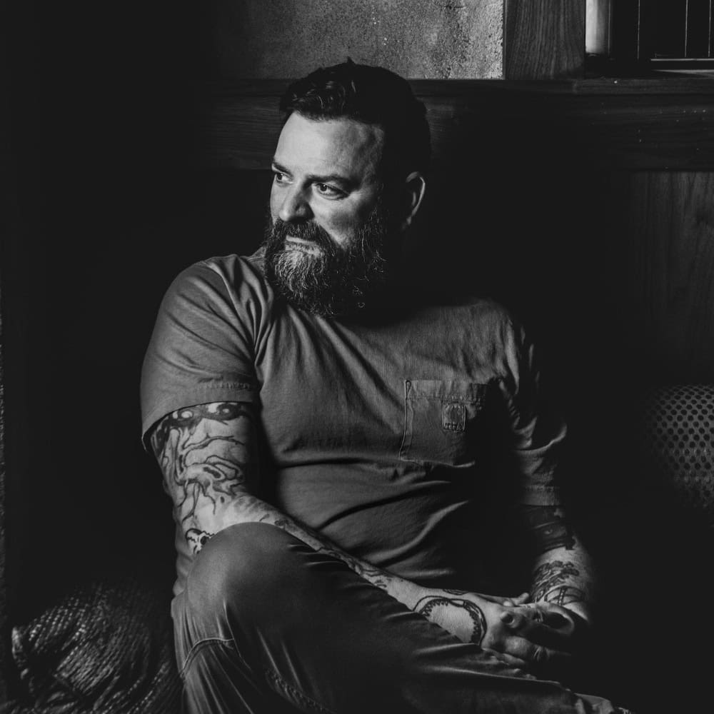 A bearded man with tattoos sits indoors in a dark, shadowy setting, looking to the side with his hands clasped.