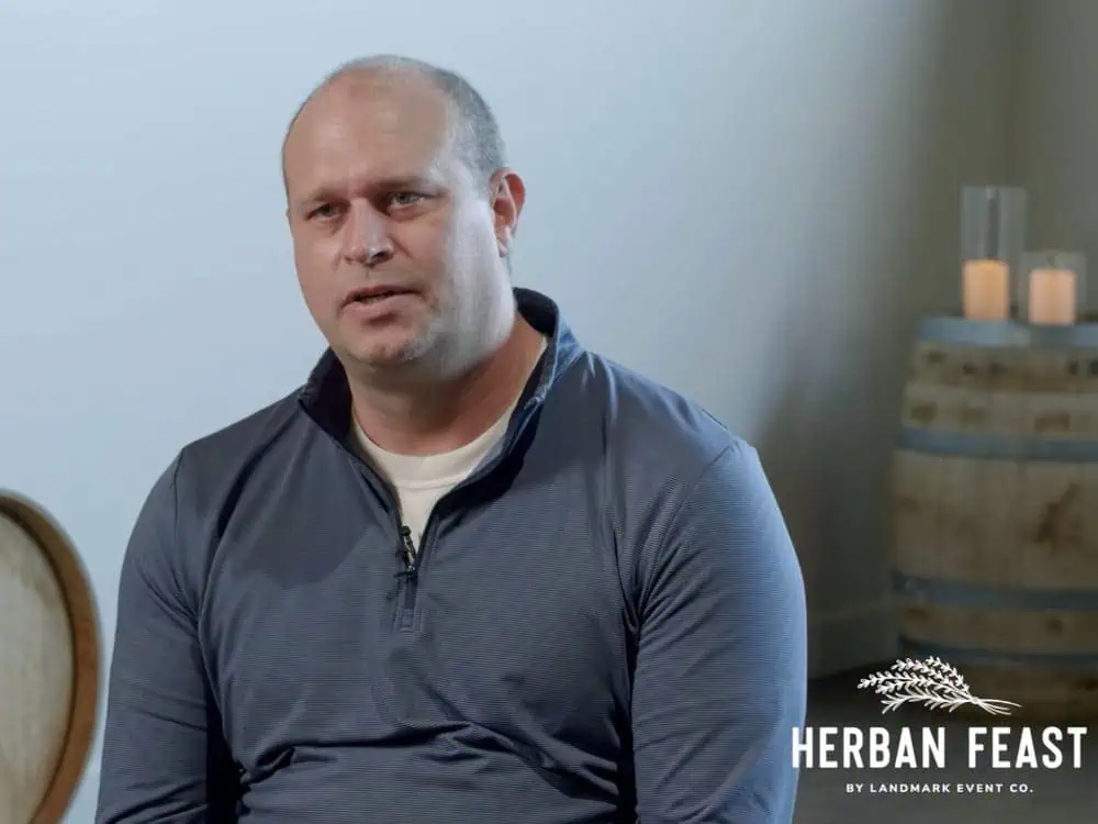 A man in a blue pullover sits indoors next to a barrel and candles, creating the perfect setting for entertaining. The “Herban Feast by Landmark Event Co.” logo appears in the lower right corner, evoking inspiration for your own holiday hosting tips.