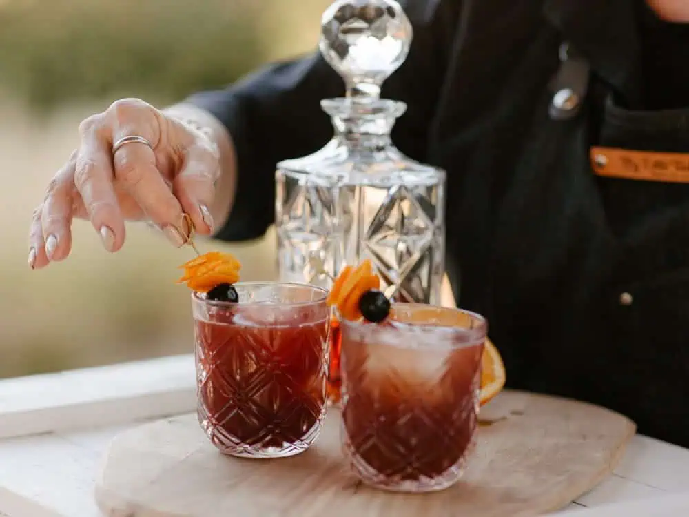 A person, perhaps taking inspiration from a culinary director at Herban Feast, places a garnish on a red cocktail in a patterned glass, with another cocktail and a crystal decanter nearby on a wooden board—a picture-perfect scene for holiday hosting tips.