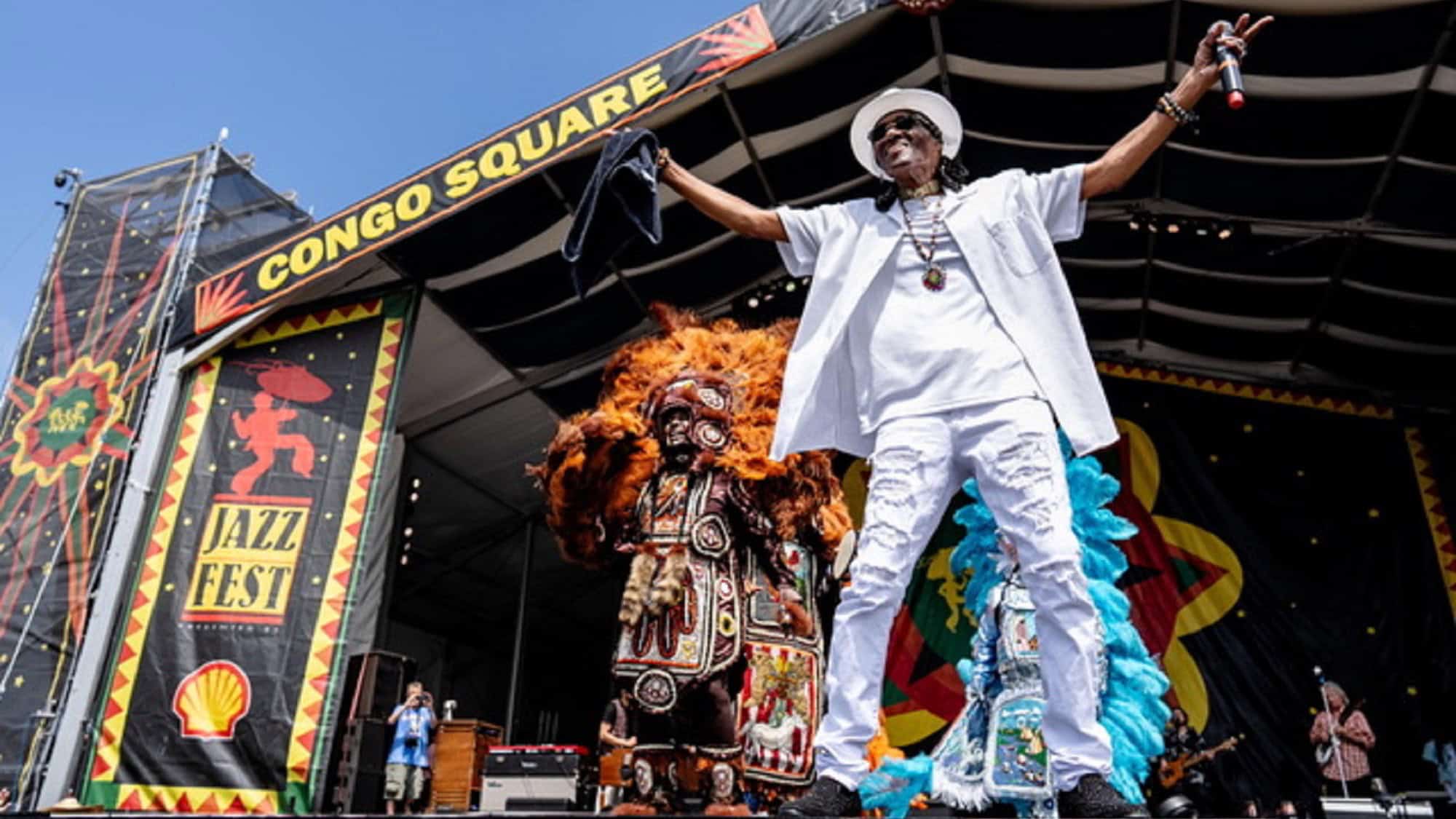 Performer in white outfit sings on stage at Jazz Fest Congo Square, next to a person in ornate, colorful costume; festival signs are visible.