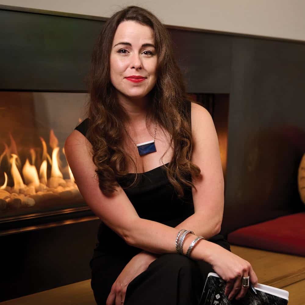 A woman with long brown hair in a black outfit sits by a fireplace, holding a book titled “Death Becomes Us” and smiling at the camera.