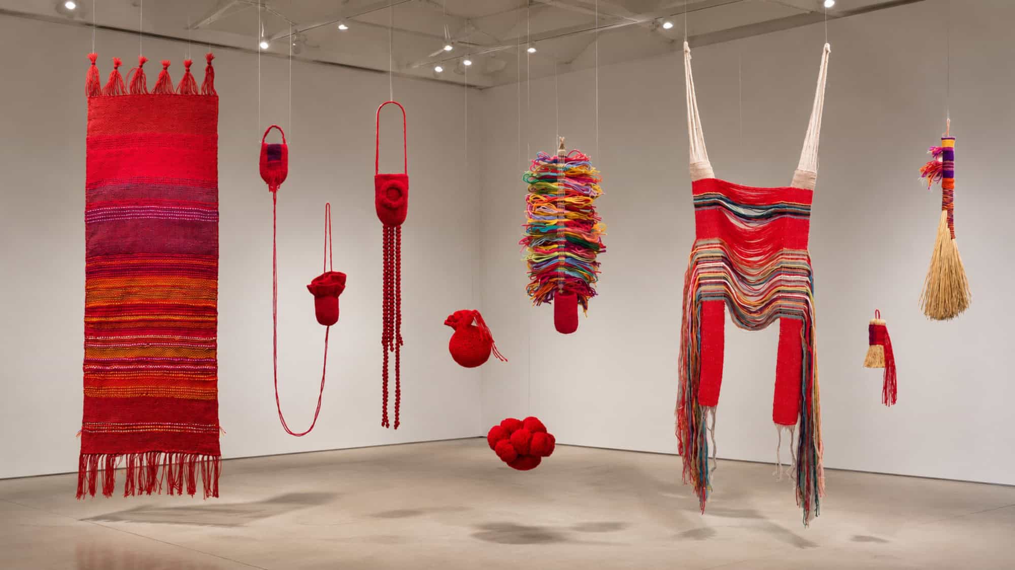 A gallery displays various red and multicolored woven and wrapped textile sculptures hanging from the ceiling, with a broom included among the pieces.