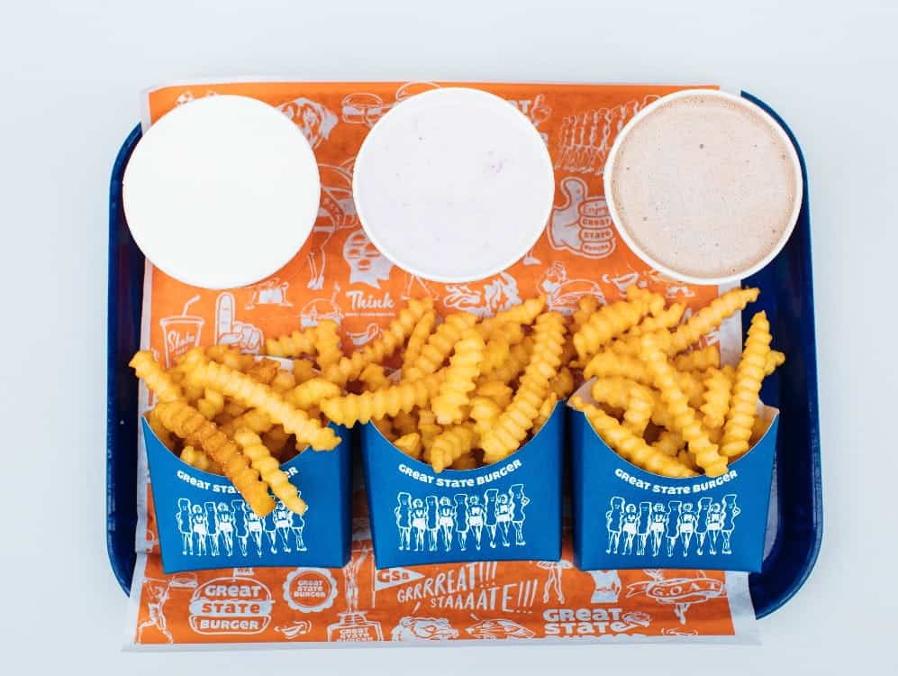 Three crinkle-cut fries portions and three drinks, including Great State Burger’s organic milkshakes, served on a blue tray with an orange paper liner featuring white graphics and text—a presentation worthy of a Seattle magazine feature.