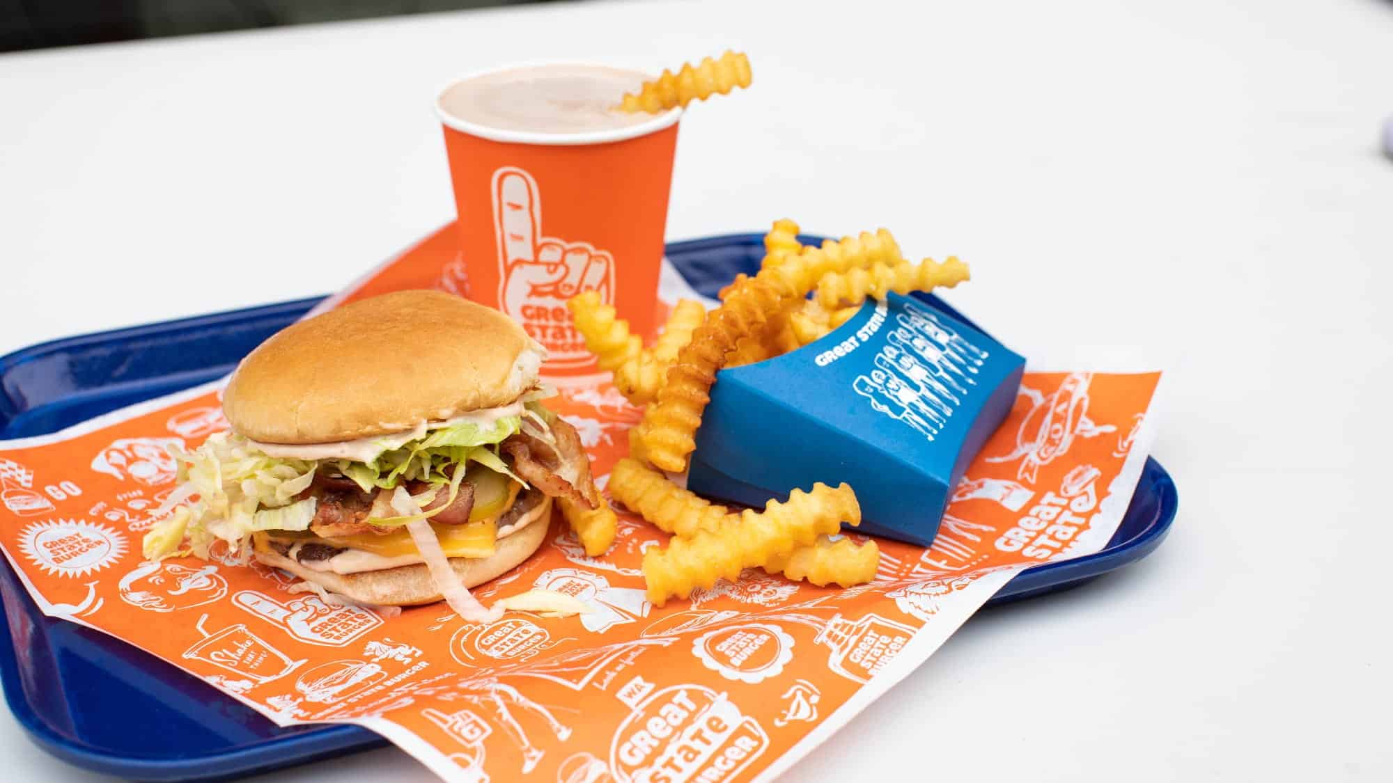 A tray with a bacon cheeseburger, crinkle-cut fries in a blue container, and a cup of soda on orange and white branded paper—just one of the delicious meals featured by Seattle magazine at Great State Burger in SeaTac Airport.