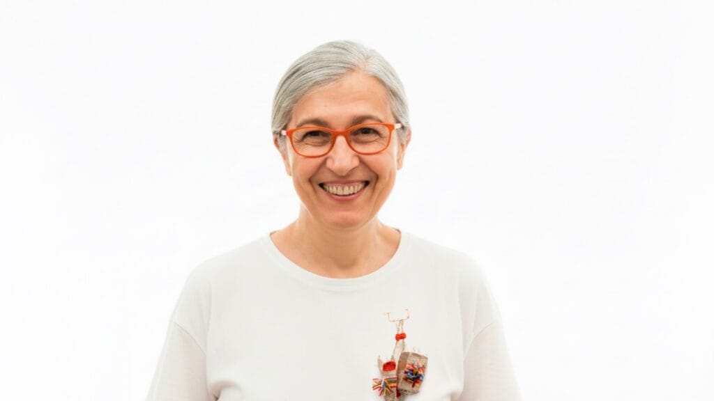 Smiling older woman with gray hair, orange glasses, and a white shirt featuring a colorful decorative brooch, standing against a white background.