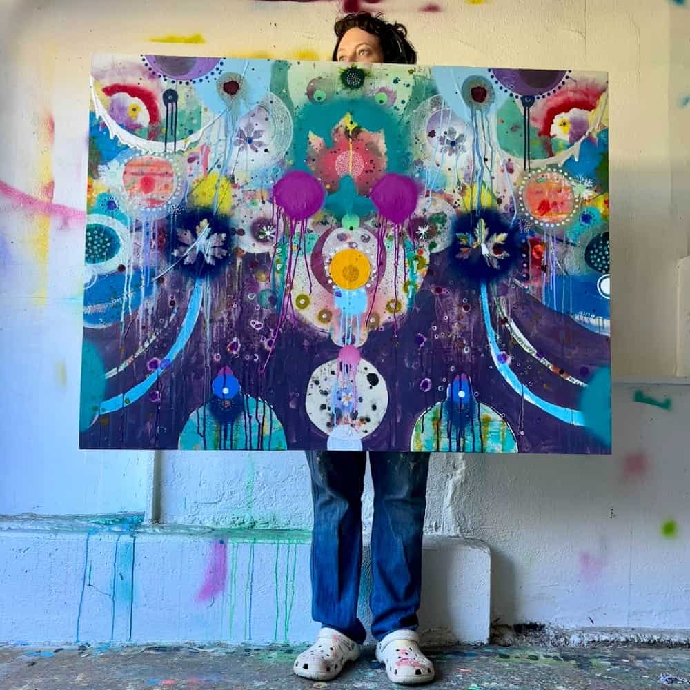 Person holding a colorful abstract painting with circles and splashes of paint, standing against a white wall with scattered paint marks—a vibrant moment from Liz Tran’s Studio Sessions. Only their jeans, shoes, and part of their head are visible.