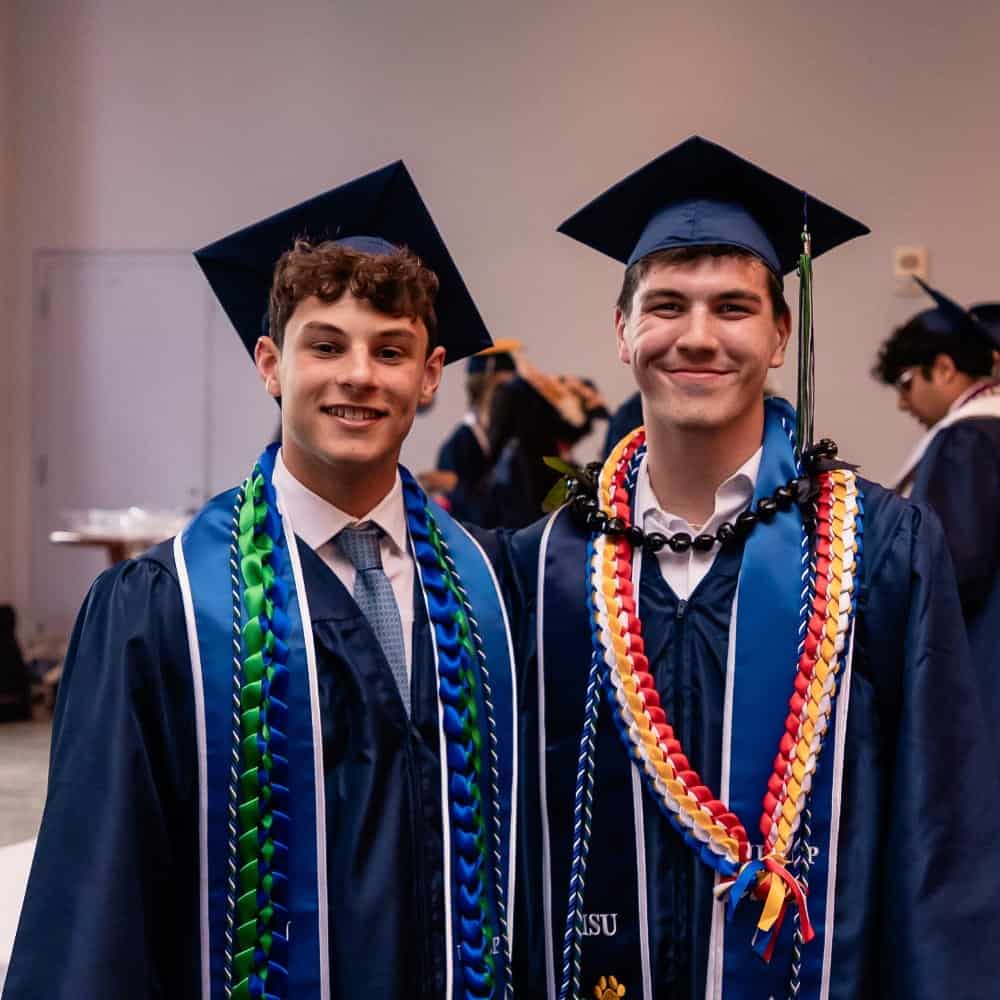 Two young men in graduation caps and gowns stand side by side, smiling. Both wear multi-colored honor cords and leis, and appear to be at an indoor graduation ceremony at UPrep in Seattle.