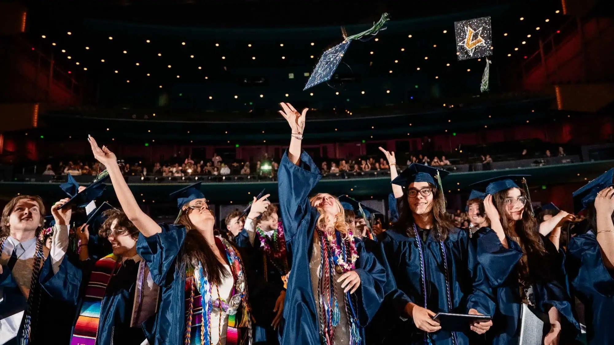 Graduates in caps and gowns celebrate by tossing caps in the air in a crowded auditorium during a graduation ceremony, marking the culmination of their hard work and benefitting from invaluable college counseling at UPrep.