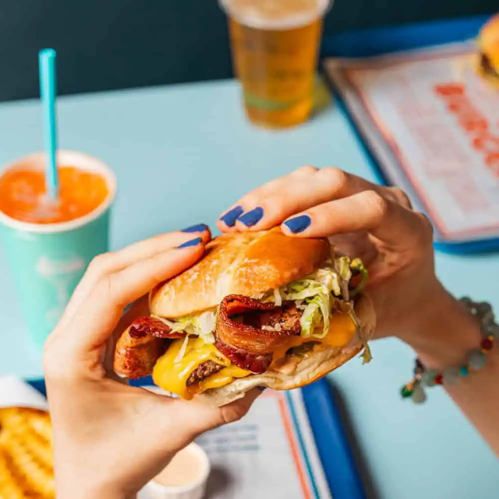 A person with blue nail polish holds a cheeseburger made with local grass-fed beef, stacked high with bacon, lettuce, and tomato. Crinkle-cut fries, a drink, and one of the airport’s signature organic milkshakes are on a tray in the background—perfect for refueling at SeaTac Airport.