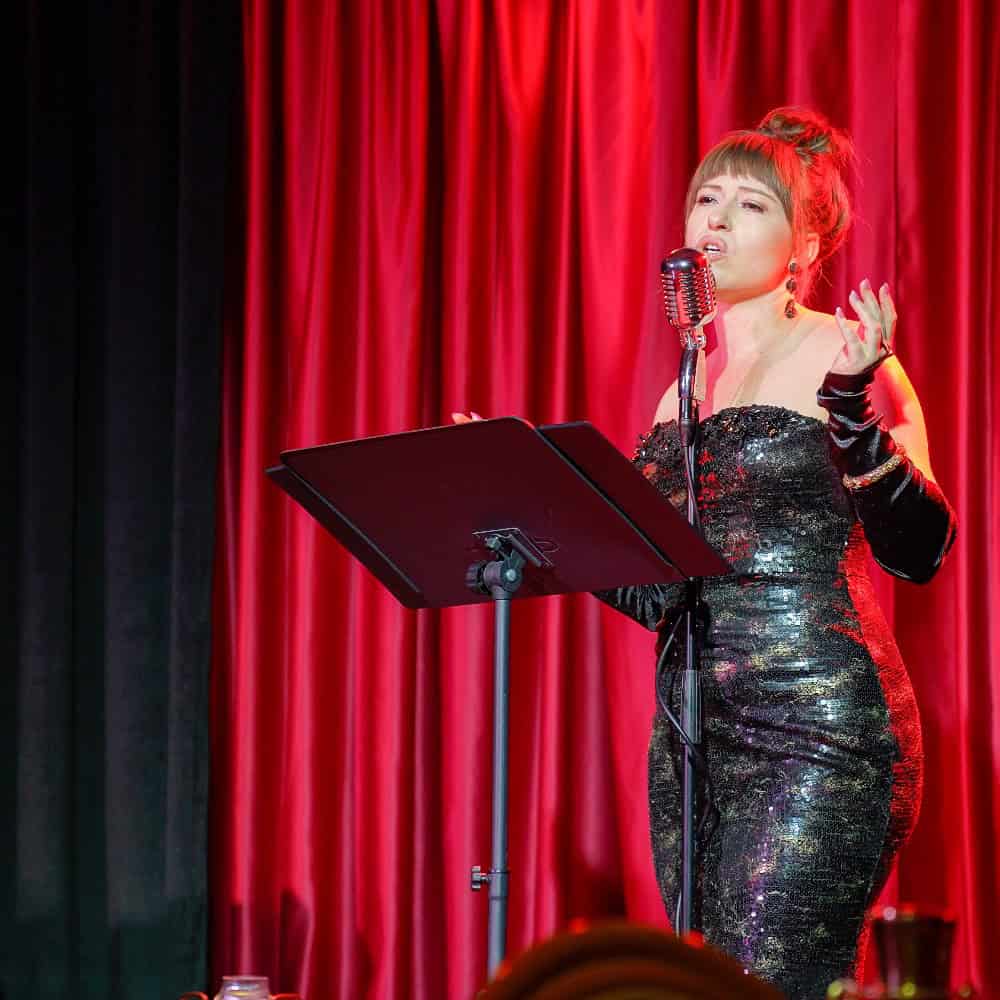 A woman in a sparkly black dress sings into a vintage microphone on stage, standing behind a music stand with red curtains in the background.