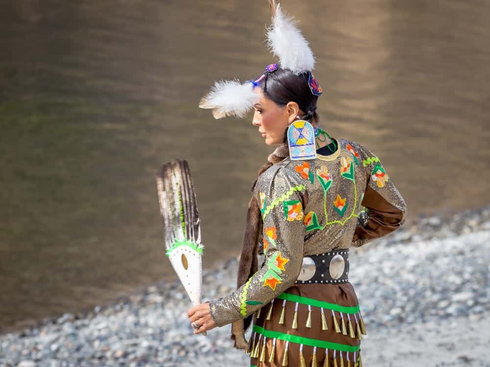 A person in traditional Native American regalia stands by a riverbank, holding a feathered fan and wearing beadwork, fringe, and feathers in their hair, embodying the rich culture and adventurous spirit of their heritage.