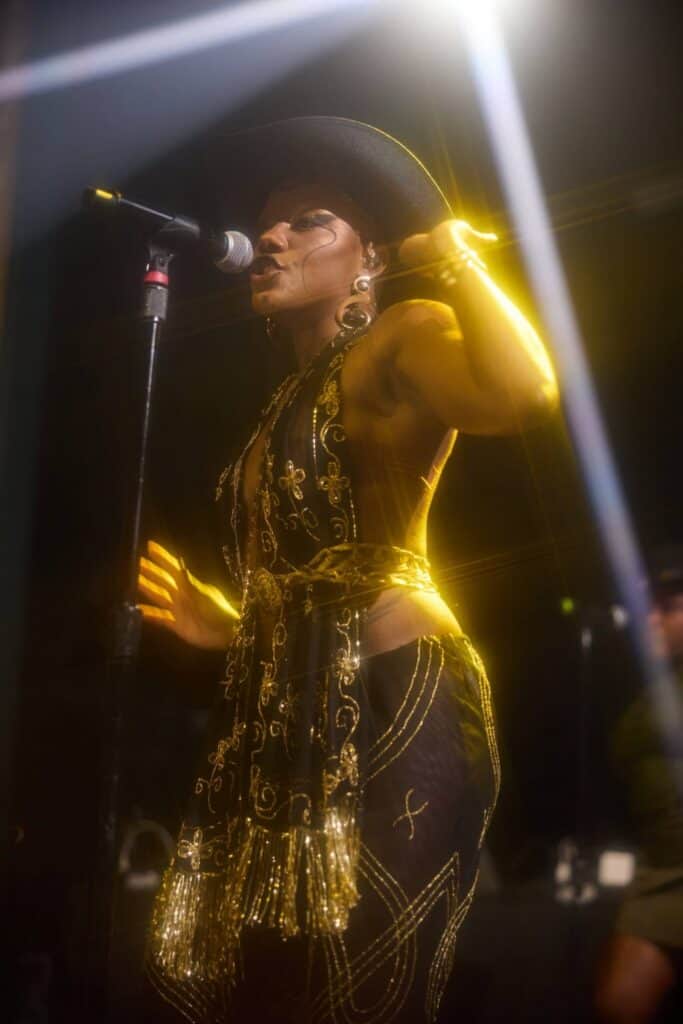 Performer in a black and gold outfit and hat sings into a microphone on stage under bright yellow stage lights.