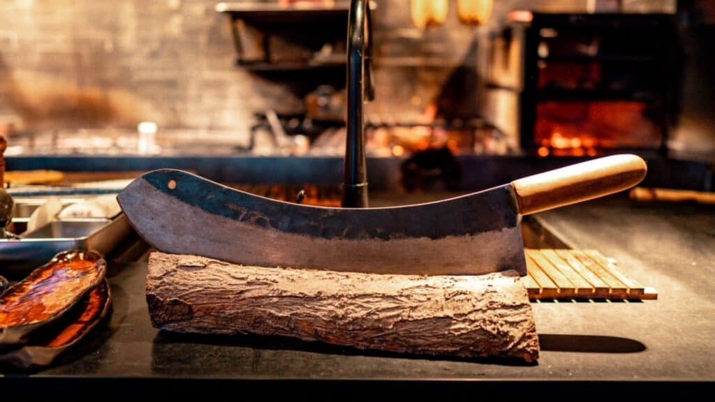 A large butcher knife with a wooden handle rests on a log on a kitchen counter, with blurred background elements and warm lighting.