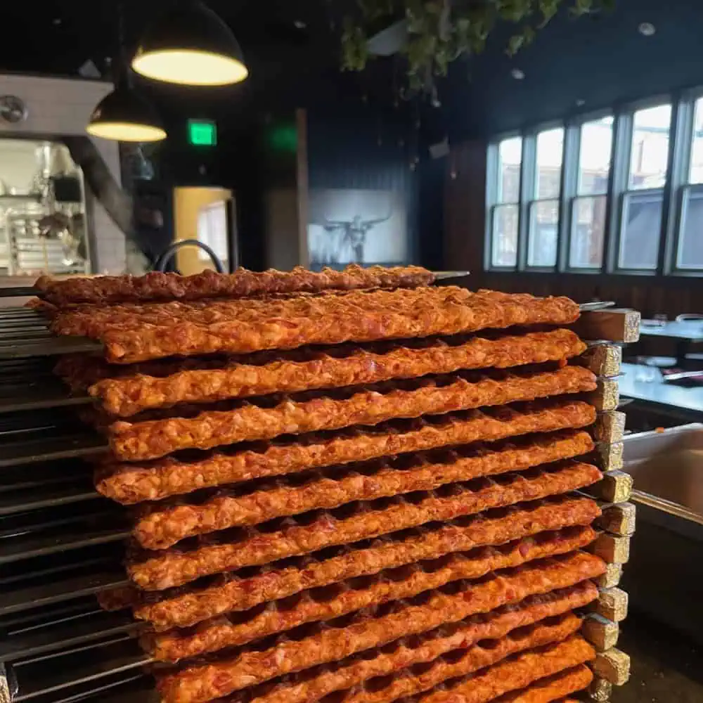 A stack of raw minced meat skewers is arranged in a restaurant kitchen with dim lighting and visible windows in the background.